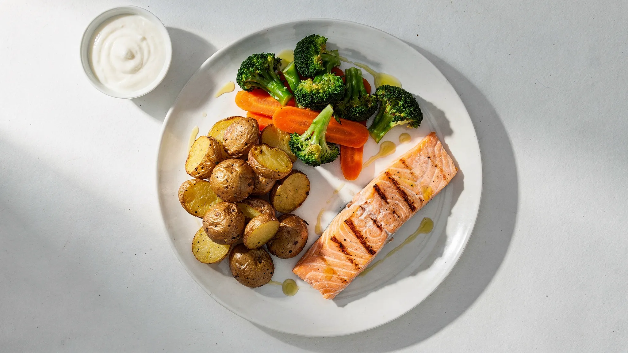 A simple balanced plate illustration using UK foods: grilled salmon, roasted new potatoes, steamed broccoli and carrots, plus a small pot of yoghurt-based sauce and a drizzle of olive oil.