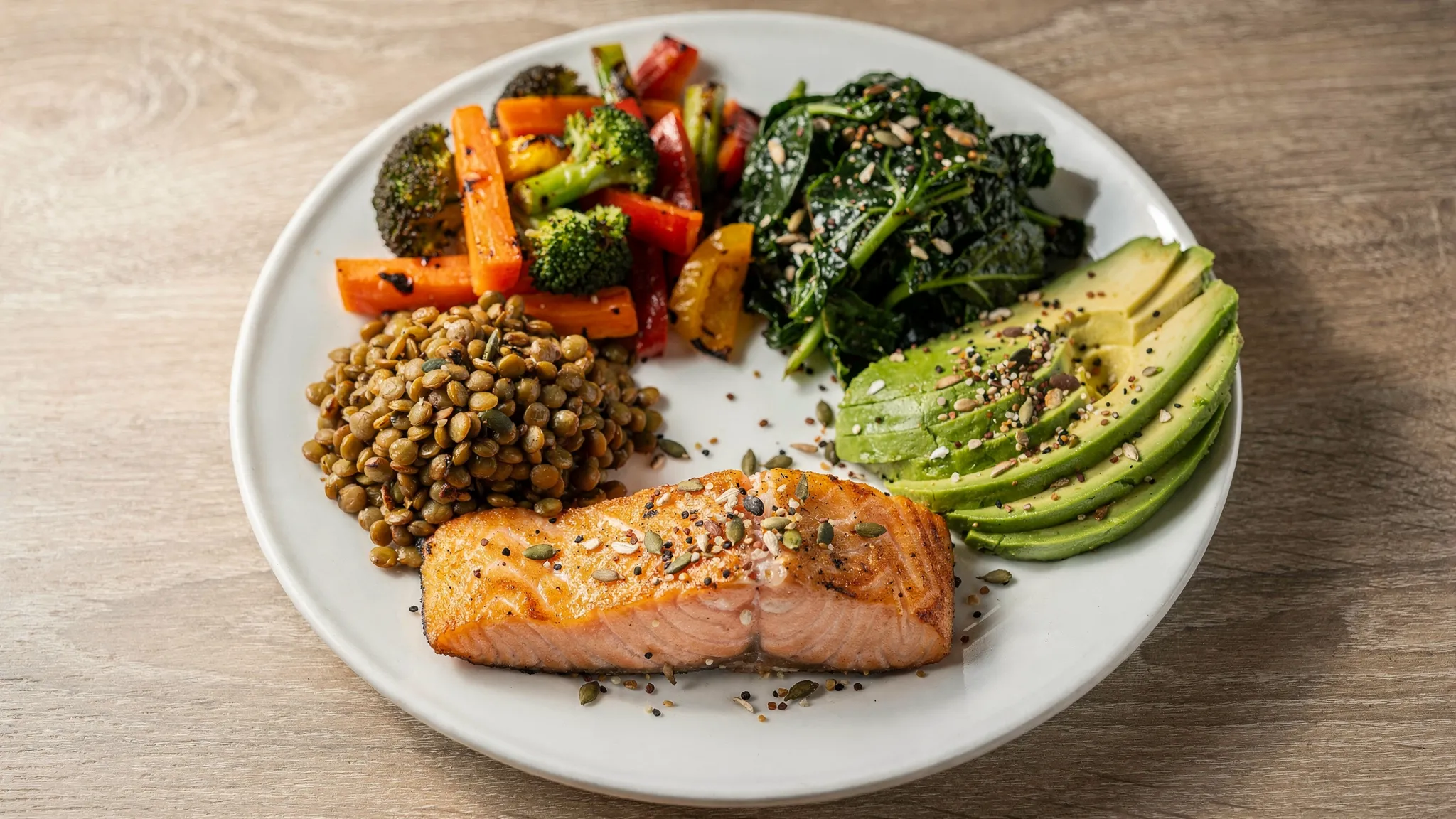 A nourishing balanced meal on a plate with salmon, roasted vegetables, lentils, leafy greens, avocado and seeds, showing protein, fibre-rich carbohydrates, colourful plants and healthy fats for satiety.